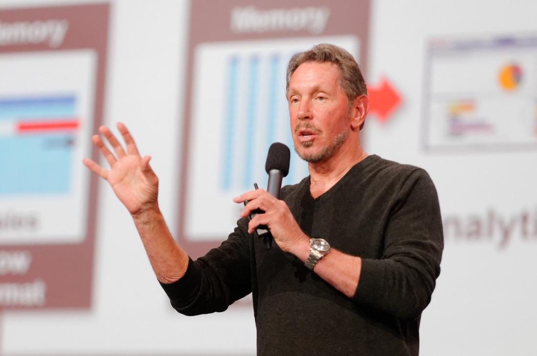 Larry Ellison on stage at Oracle OpenWorld in 2013, microphone in hand, in front of a slide