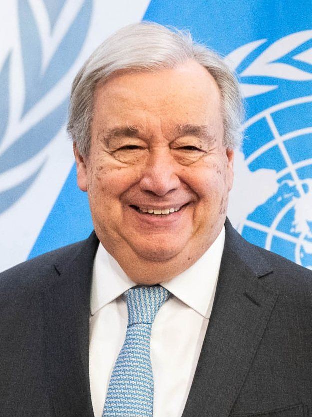 Smiling man in suit, UN emblem and flag visible in background.
