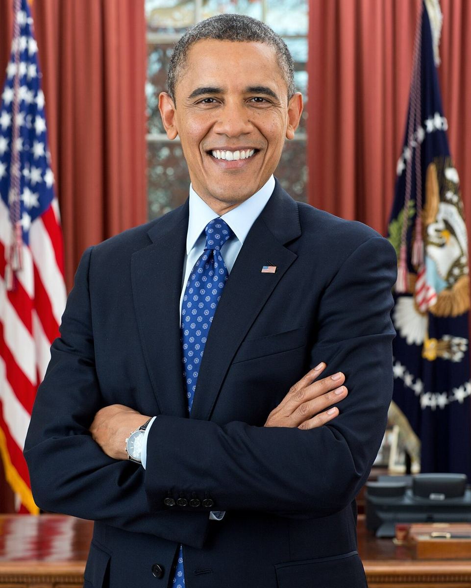 smiling man in suit, posed official portrait with flags behind