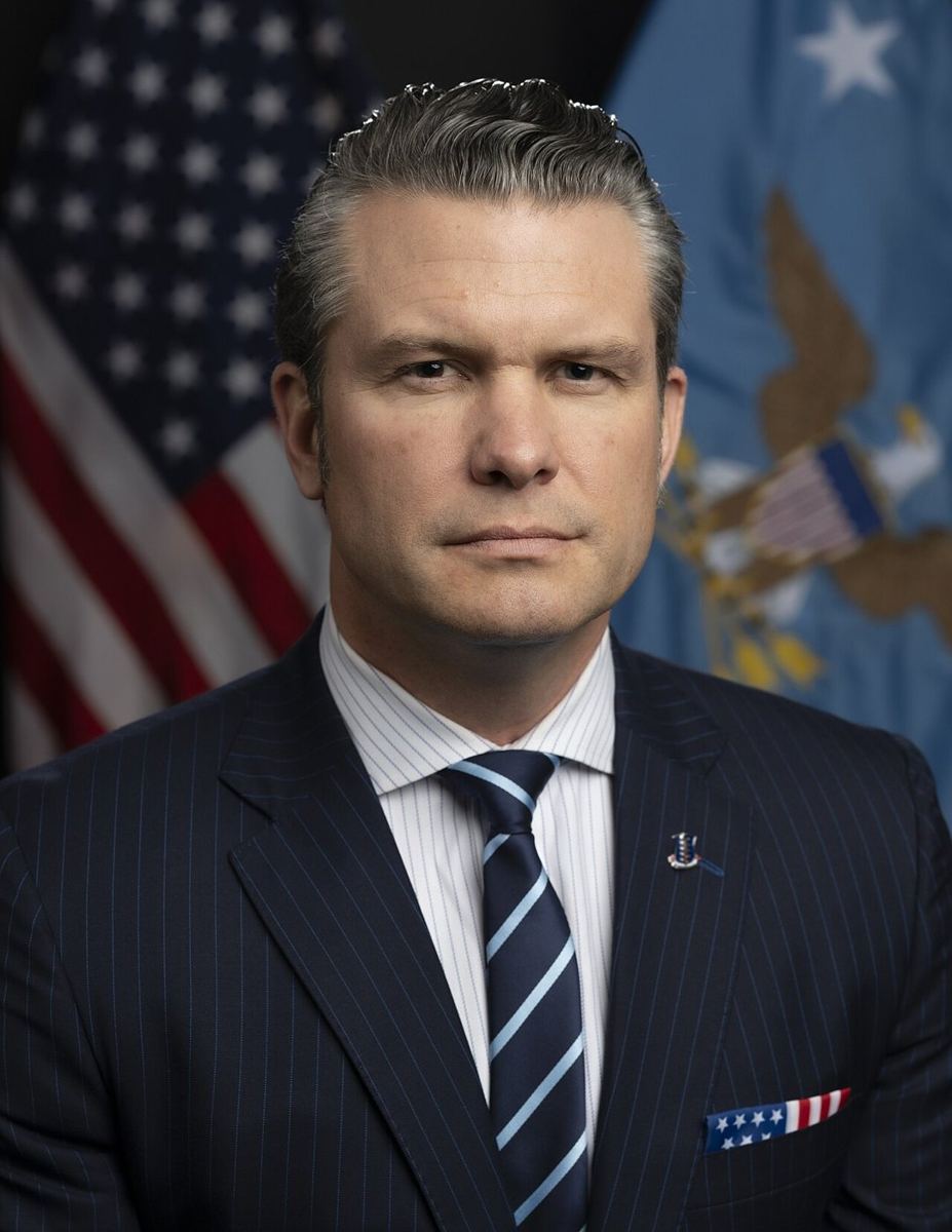 Head-and-shoulders portrait of a man in suit, flags behind.